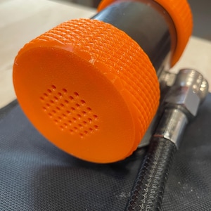 May include: Close-up of an orange and black nozzle with a textured grip. The nozzle has a series of small holes and is connected to a black hose with a silver fitting. The object is on a dark surface.