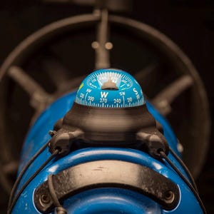 May include: A blue and black compass mounted on a blue object. The compass has a blue face with white and black markings. The background includes a dark wheel and other dark objects.