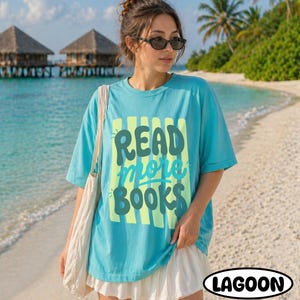 Puede incluir: Una camiseta turquesa con las palabras "READ more BOOKS" en un dise&ntilde;o verde y amarillo. La camiseta se usa en una playa con una falda blanca y un bolso de mano blanco. La palabra "LAGOON" est&aacute; en un &oacute;valo negro.