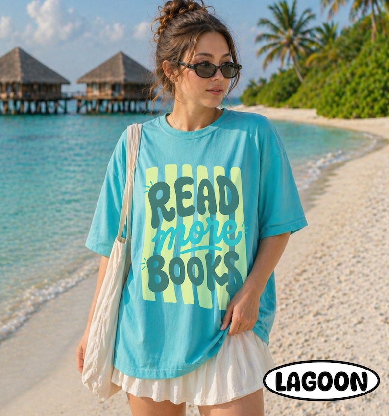 Puede incluir: Una camiseta turquesa con las palabras "READ more BOOKS" en un dise&ntilde;o verde y amarillo. La camiseta se usa en una playa con una falda blanca y un bolso de mano blanco. La palabra "LAGOON" est&aacute; en un &oacute;valo negro.