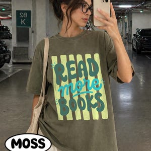 Puede incluir: Camiseta verde oliva con el texto "READ more BOOKS" en dise&ntilde;o turquesa y amarillo. La camiseta tiene un corte holgado y mangas cortas. Se ve un bolso tote blanco sobre el hombro. La palabra "MOSS" est&aacute; en un &oacute;valo blanco en la parte inferior.