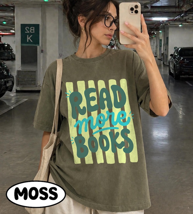 Puede incluir: Camiseta verde oliva con el texto "READ more BOOKS" en dise&ntilde;o turquesa y amarillo. La camiseta tiene un corte holgado y mangas cortas. Se ve un bolso tote blanco sobre el hombro. La palabra "MOSS" est&aacute; en un &oacute;valo blanco en la parte inferior.