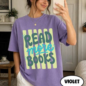 Puede incluir: Camiseta violeta con el texto "READ more BOOKS" en verde y azul. El texto est&aacute; sobre un fondo de rayas verdes verticales. La camiseta es de corte holgado y parece estar hecha de material suave.