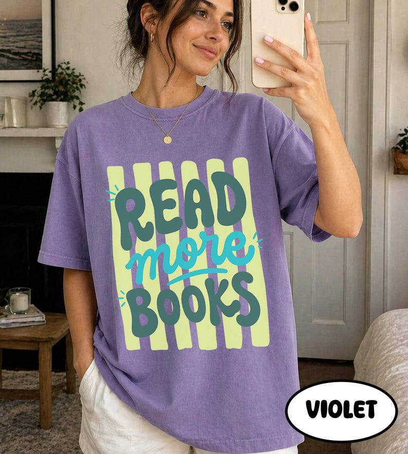 Puede incluir: Camiseta violeta con el texto "READ more BOOKS" en verde y azul. El texto est&aacute; sobre un fondo de rayas verdes verticales. La camiseta es de corte holgado y parece estar hecha de material suave.