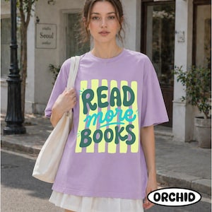 Puede incluir: Una camiseta lila con la frase "READ more BOOKS" en verde y turquesa sobre un fondo de rayas amarillas. La camiseta forma parte de un atuendo informal. La palabra "ORCHID" est&aacute; impresa en la parte inferior derecha.
