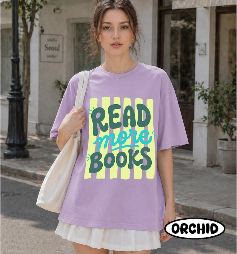 Puede incluir: Una camiseta lila con la frase "READ more BOOKS" en verde y turquesa sobre un fondo de rayas amarillas. La camiseta forma parte de un atuendo informal. La palabra "ORCHID" est&aacute; impresa en la parte inferior derecha.