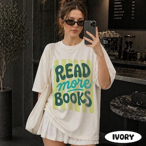 Puede incluir: Camiseta de color marfil con el texto "READ more BOOKS" en verde y turquesa. La camiseta tiene un corte holgado y mangas cortas. Tambi&eacute;n se ve un bolso tote blanco y una falda blanca.