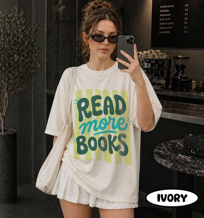 Puede incluir: Camiseta de color marfil con el texto "READ more BOOKS" en verde y turquesa. La camiseta tiene un corte holgado y mangas cortas. Tambi&eacute;n se ve un bolso tote blanco y una falda blanca.