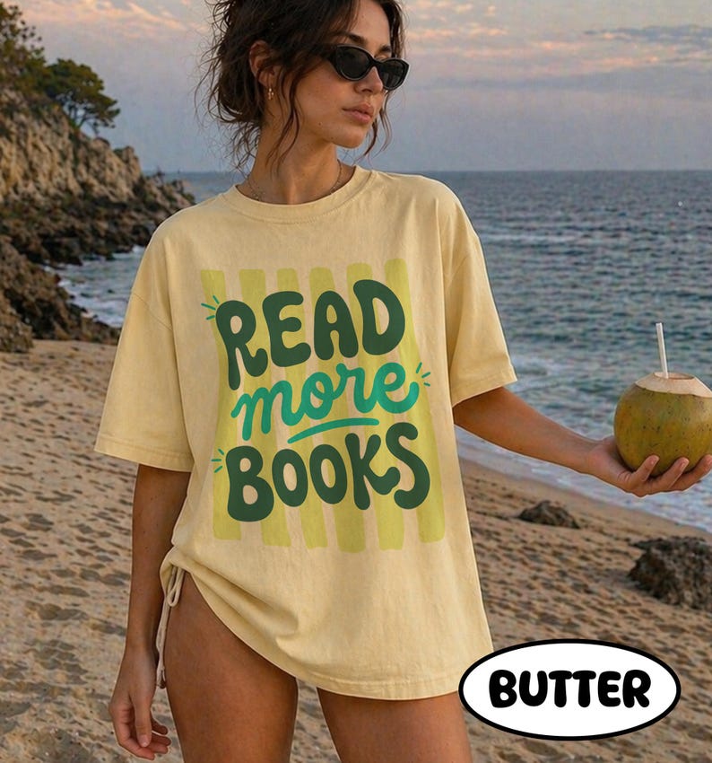 Puede incluir: Camiseta de color mantequilla con el texto "READ more BOOKS" en verde y turquesa. La camiseta tiene un corte holgado y se lleva en la playa. Una persona sostiene un coco con una pajita.