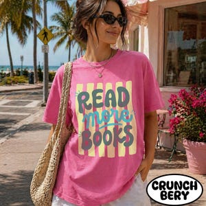 Puede incluir: Una camiseta rosa con el texto "READ more BOOKS" en un dise&ntilde;o colorido. La camiseta se combina con pantalones cortos blancos y una bolsa tejida. El fondo incluye palmeras y una se&ntilde;al de tr&aacute;fico.