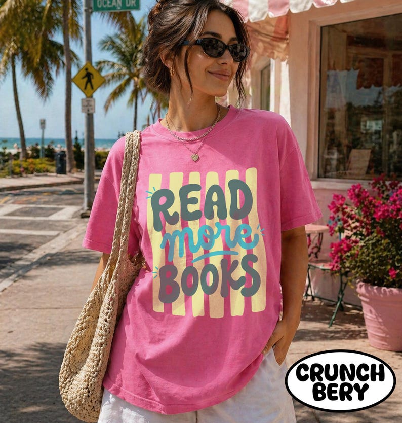 Puede incluir: Una camiseta rosa con el texto "READ more BOOKS" en un dise&ntilde;o colorido. La camiseta se combina con pantalones cortos blancos y una bolsa tejida. El fondo incluye palmeras y una se&ntilde;al de tr&aacute;fico.