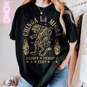 May include: Black t-shirt with a gold tiger graphic and the words "CHINGA LA MIGRA" above the tiger. Below the tiger are the words "RESIST PERSIST EXIST" with star accents. The shirt is a comfort color.