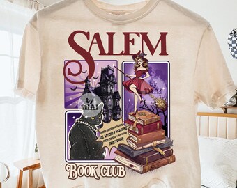 May include: Cream-colored t-shirt featuring a Salem Book Club design. The graphic includes a black cat, a witch, a haunted house, and stacked books. Text on the shirt reads "Salem Book Club" and "All Witches Welcome Every Tuesday & Saturday in October."