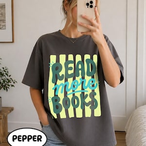 Puede incluir: Camiseta gris oscuro con el texto "READ more BOOKS" en un dise&ntilde;o verde y azul. El texto est&aacute; sobre un fondo a rayas amarillas. La palabra "PEPPER" est&aacute; en un &oacute;valo blanco en la parte inferior.