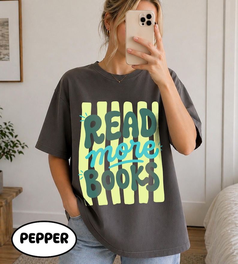 Puede incluir: Camiseta gris oscuro con el texto "READ more BOOKS" en un dise&ntilde;o verde y azul. El texto est&aacute; sobre un fondo a rayas amarillas. La palabra "PEPPER" est&aacute; en un &oacute;valo blanco en la parte inferior.