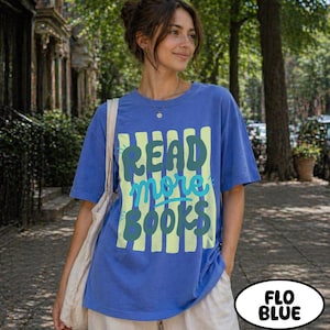 Puede incluir: Una camiseta azul con el texto "READ more BOOKS" en verde y azul claro. El texto est&aacute; sobre un fondo de rayas verticales amarillas. La camiseta es de color azul liso y tiene mangas cortas.