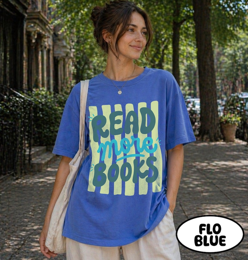 Puede incluir: Una camiseta azul con el texto "READ more BOOKS" en verde y azul claro. El texto est&aacute; sobre un fondo de rayas verticales amarillas. La camiseta es de color azul liso y tiene mangas cortas.
