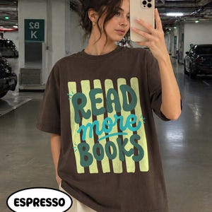 Puede incluir: Camiseta marr&oacute;n con la frase "READ more BOOKS" en verde y turquesa. El texto est&aacute; sobre un fondo de rayas verticales verde claro. La camiseta es de estilo casual y de color 'espresso'.