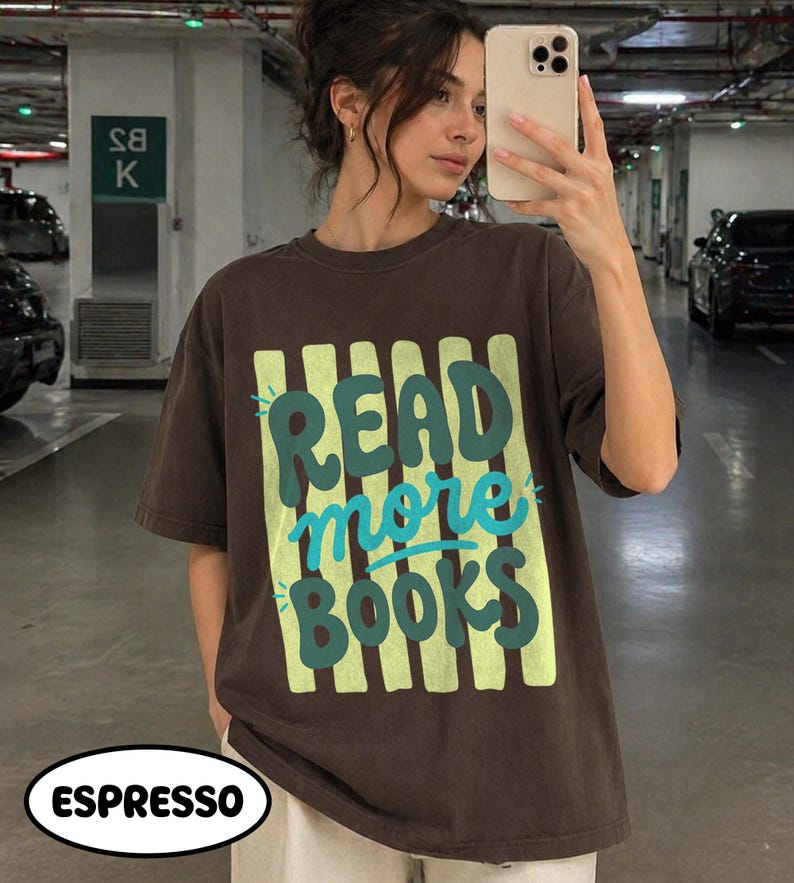 Puede incluir: Camiseta marr&oacute;n con la frase "READ more BOOKS" en verde y turquesa. El texto est&aacute; sobre un fondo de rayas verticales verde claro. La camiseta es de estilo casual y de color 'espresso'.