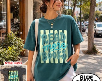May include: A dark teal short-sleeve t-shirt featuring a graphic print of vertical light green stripes and the text "READ more BOOKS" in a bold, rounded teal font. The shirt has a crew neckline and a relaxed, loose-fitting silhouette.