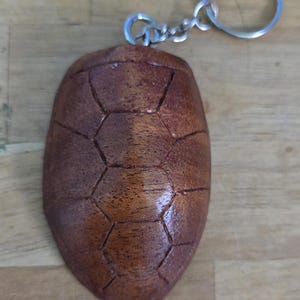 May include: A handcrafted wooden turtle shell keychain. The keychain features a detailed, carved turtle shell design with a natural wood grain finish. The keychain is attached to a silver-colored metal ring and chain.