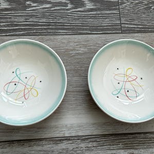 May include: Two round ceramic bowls with a white interior and a light blue rim. The center of each bowl features a colorful graphic design consisting of thin, intersecting lines in yellow, pink, and blue, accented by small black star-shaped symbols.