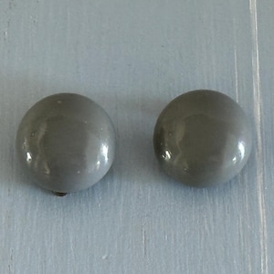 May include: Two gray, round, button-shaped earrings with a glossy finish. The earrings are set against a light blue, textured surface. The earrings are a simple, classic design.