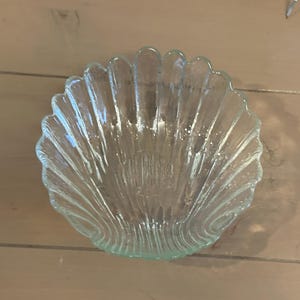 May include: A clear glass bowl shaped like a seashell, featuring a scalloped edge and vertical ridges that radiate from the center to the rim, creating a textured, translucent appearance with a smooth, concave interior surface.
