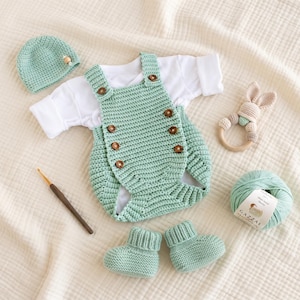 May include: A mint green crochet romper with brown buttons, a matching beanie, and booties. Includes a white long-sleeve shirt, a wooden crochet hook, a beige bunny rattle, and a ball of mint green yarn with a label reading Gazzal.