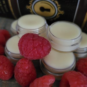 Könnte beinhalten: Nahaufnahme von mehreren kleinen, durchsichtigen Behältern, gefüllt mit weißem Lippenbalsam, umgeben von roten Himbeeren. Die Behälter haben einen goldenen Deckel mit einem Schlüsselloch-Design.