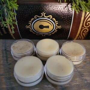 May include: Five small, clear jars of lip balm are arranged in a semi-circle. The lip balms are light beige and tan in color. The background includes a decorative box with gold accents and the word "HOLMES".