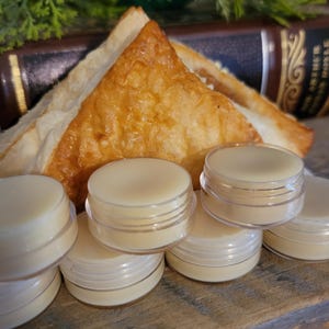 May include: A collection of small, clear plastic containers filled with a light yellow balm. The containers are stacked on a wooden surface, with a golden-brown pastry in the background. Green foliage and a dark book are also visible.