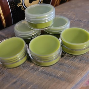 May include: Six small clear plastic containers filled with a bright green balm. The containers are arranged in a semi-circle on a wooden surface.