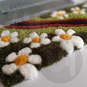 May include: Close-up of a handmade rug with a daisy pattern. The rug features white daisy flowers with yellow centers on a green and brown background. The word "TUFTRUGS" is visible in the bottom right corner.