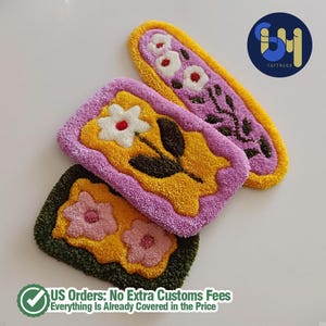 May include: Three colorful, tufted rugs with floral designs. One is rectangular with a white flower, yellow center, and purple border. Another is oval with white flowers and a purple floral design. The third is square with pink flowers.