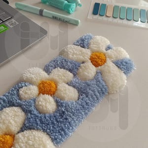 May include: A handmade, rectangular rug with a daisy flower design. The rug features white, yellow, and light blue tufted yarn. The text "TUFTRUGS" is visible on the bottom right corner. The rug is on a white surface.