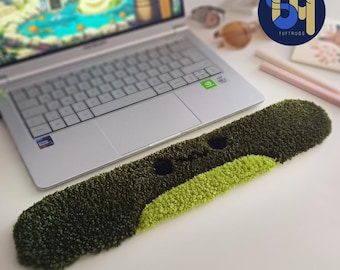 May include: A green and black tufted rug with a smiling face design, placed in front of a laptop. The rug is rectangular and has a textured appearance. The laptop is silver and open, displaying a video game.