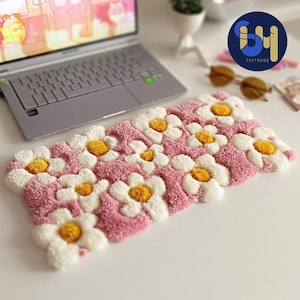 May include: A rectangular tufted rug featuring a repeating pattern of white daisy-like flowers with bright yellow centers, set against a textured pink background. The rug has a thick, plush pile and a soft, looped surface texture throughout.