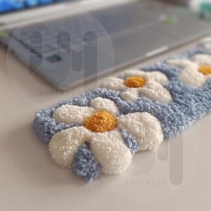 May include: A blue and white daisy-patterned rug with yellow centers. The rug is rectangular and has a textured, tufted appearance. The background is blurred, with a laptop visible.