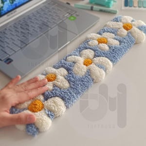 May include: A blue and white daisy-patterned rug, with yellow centers, rests on a white surface. The rug is rectangular and has a fluffy texture. The word "TUFTRUGS" is visible on the rug. A laptop is in the background.