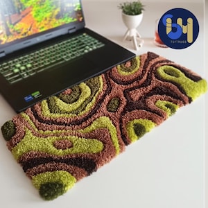 Peut inclure: Un tapis de couleur verte, marron et vert foncé à motifs abstraits avec un ordinateur portable posé dessus. Le tapis est fait de fil touffeté et présente un design ondulé et organique.