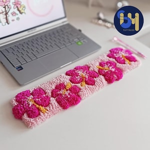 May include: A rectangular tufted keyboard wrist rest featuring a light pink background with a repeating pattern of vibrant magenta hibiscus flowers. Each flower has a bright yellow center and is crafted with a textured, plush yarn pile surface.