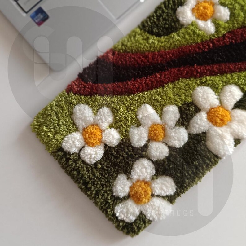 May include: A handmade, rectangular rug featuring a floral design. The rug has a green background with white daisy-like flowers with yellow centers. A dark red and black stripe runs across the top. The rug is likely made of a soft, textured material.