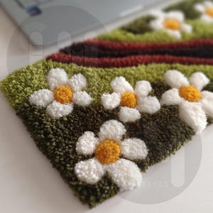 May include: Close-up of a handmade rug featuring a daisy design. The rug has a green and brown background with white daisies and yellow centers. A red stripe is visible near the top. The rug is made of a textured, looped material.