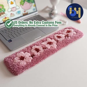 May include: A rectangular, pink, tufted rug with a floral pattern. The rug features five flowers with dark red centers and lighter pink petals. The text "US Orders: No Extra Customs Fees Everything Is Already Covered in the Price" is visible. The rug is placed near a laptop.