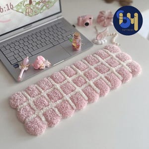 May include: A rectangular tufted keyboard wrist rest featuring a grid pattern of soft pink plush squares separated by white lines. The textured, cushioned accessory is positioned on a flat surface next to a silver laptop and various small decorative items.