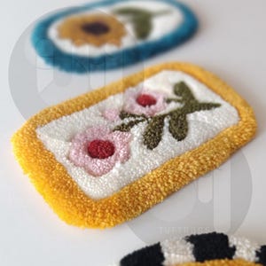 May include: A yellow framed rug with a white background and two pink flowers with red centers and green leaves. The rug is made with a fluffy texture.