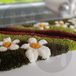 May include: Close-up of a daisy-themed rug with white petals and yellow centers. The rug features green, brown, and red stripes, with the text "TUFTRUGS" visible in the bottom right corner. The rug is likely handmade.