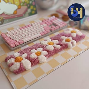 May include: A pink and white mechanical keyboard with a matching pink and white daisy-shaped wrist rest. The wrist rest has white and yellow flowers. The keyboard and wrist rest are on a yellow and white checkered mousepad.