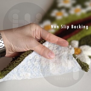 May include: Close-up of a rug with a white, non-slip backing. The rug has a green border and a floral design with white and yellow flowers. The text "Non Slip Backing" is visible.